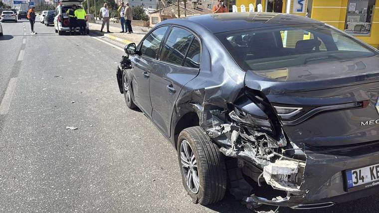Yol kenarında duran gri otomobilin arka kısmı ağır hasarlı; uzakta birkaç kişi ve başka araçlar görülüyor.