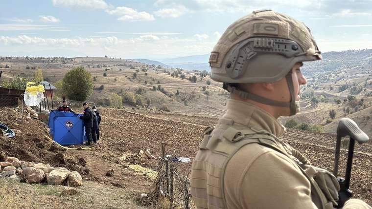 Askeri bir personel, arazide mavi bir çadırın önünde duruyor; arka planda dağlık bir manzara var.