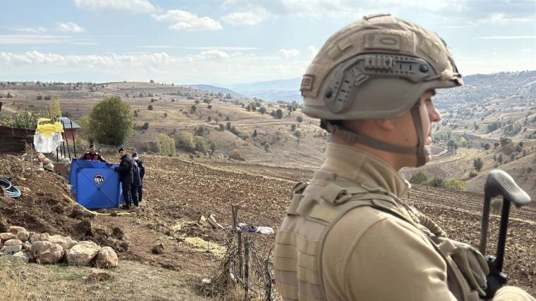 Askeri bir personel, arazide mavi bir çadırın önünde duruyor; arka planda dağlık bir manzara var.