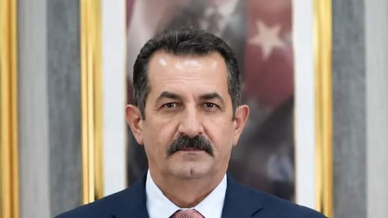 Kürsüde konuşan bir kişi, arka planda Türk bayrağı ve çerçeveli bir portre var.