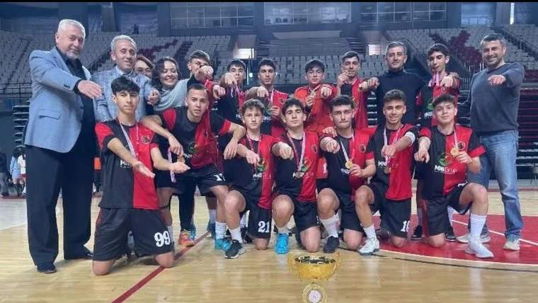 Kırmızı-siyah formalar giyen futsal oyuncuları, salonda kupa ve madalyalarla birlikte poz veriyor.