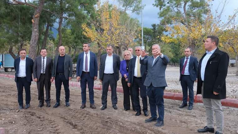 Bir grup insan, ağaçların arasında bir alanda duruyor ve bir kişi el işaretiyle konuşuyor.