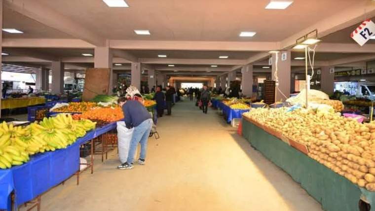Kapalı pazar alanında meyve ve sebze tezgâhları arasında alışveriş yapan insanlar görülüyor.