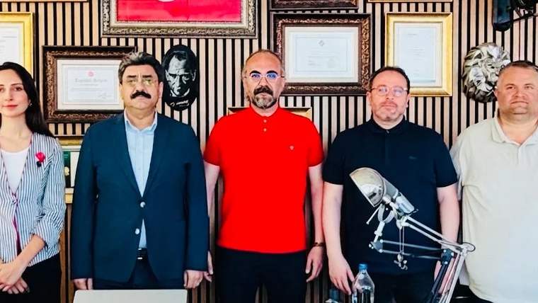 Üç kişi, bir odada yan yana duruyor; arka planda çerçevelenmiş belgeler ve tablolar var.