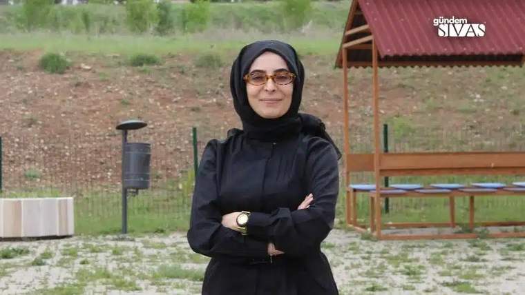 Açık alanda siyah başörtülü ve gözlüklü bir kadın, kollarını göğsünde kavuşturmuş duruyor.