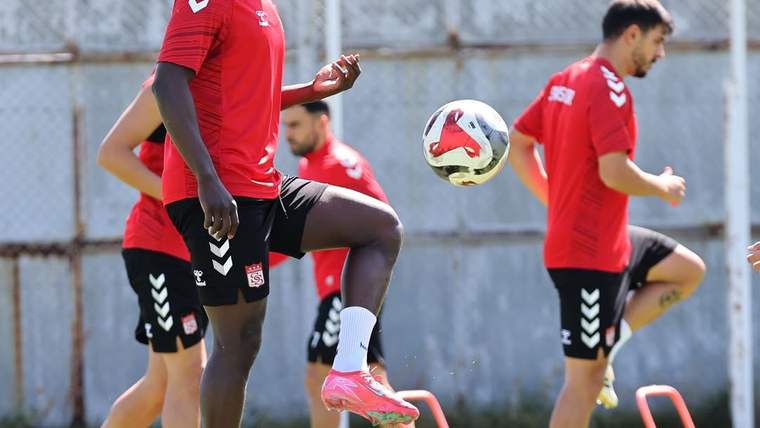 Kırmızı formalı futbolcular, antrenman sahasında havadaki top ve alçak engellerle çalışma yapıyor.
