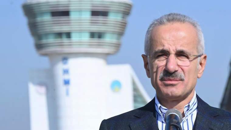 Mikrofon önünde konuşan bıyıklı bir kişi, arka planda havalimanı kontrol kulesiyle görülüyor.