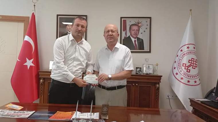 Bir odada iki kişi, Türk bayrağı ve kurum flamalarının önünde birlikte bir belge tutuyor.