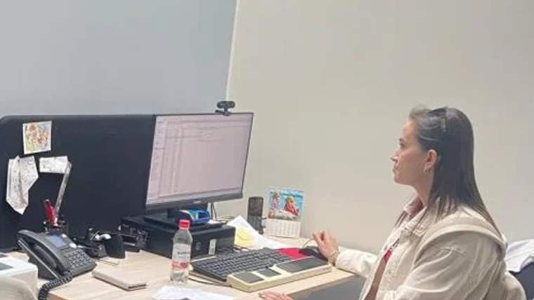 Bir kadın, bilgisayar başında çalışıyor; masada telefon, takvim ve su şişesi var.