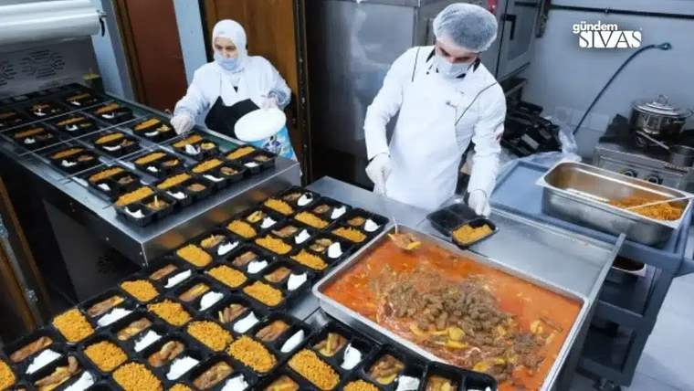 Bir mutfakta, yemekleri porsiyonlayan iki kişi, masada yemek kaplarıyla çalışıyor.