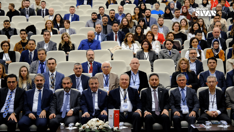 Finans sempozyumuna katılan kalabalık bir izleyici grubu, sahnede oturan konuşmacılarla birlikte görüntüleniyor.