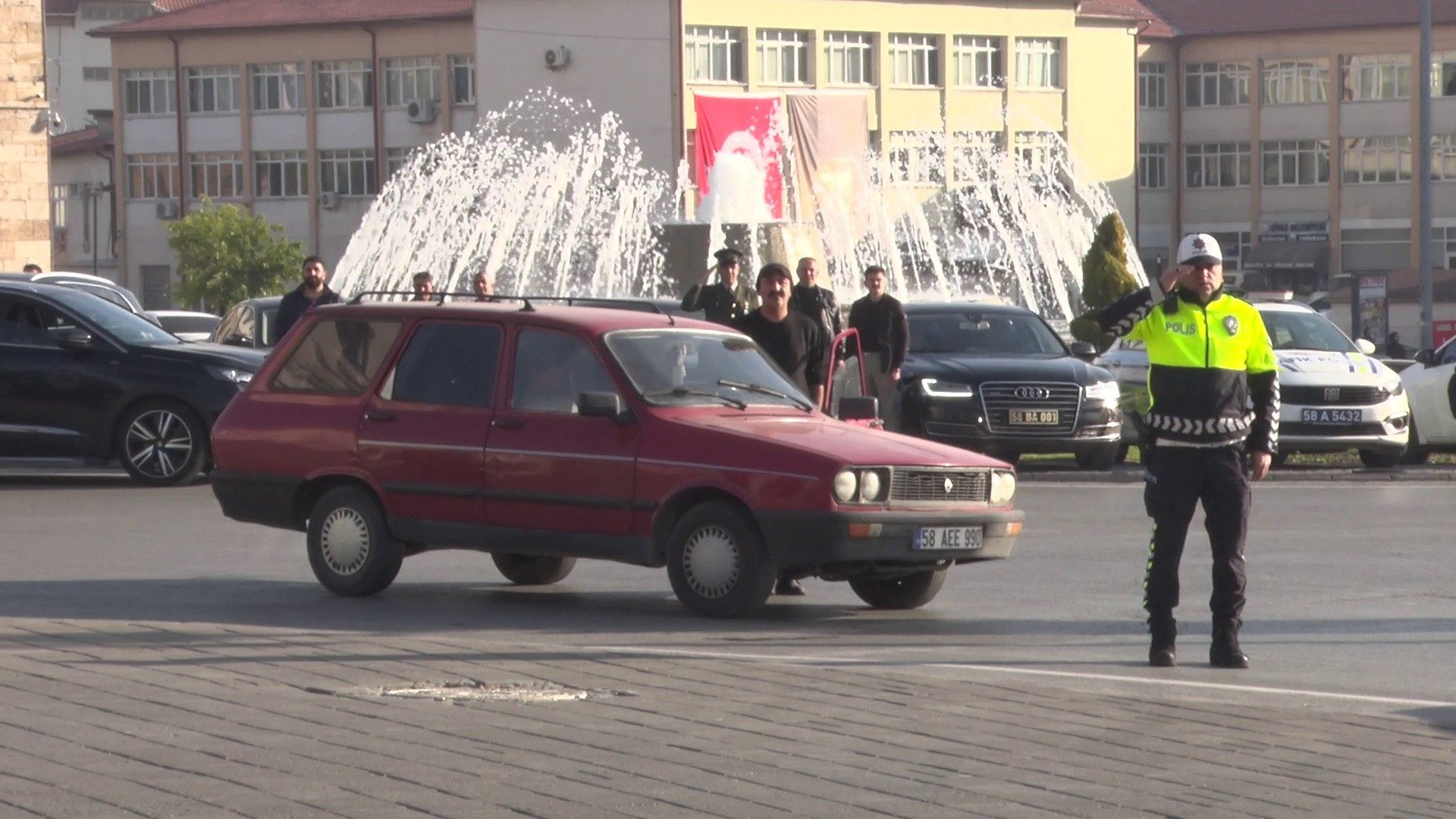 Kırmızı bir otomobil, polis memurunun yönlendirmesiyle bir meydanda duruyor.