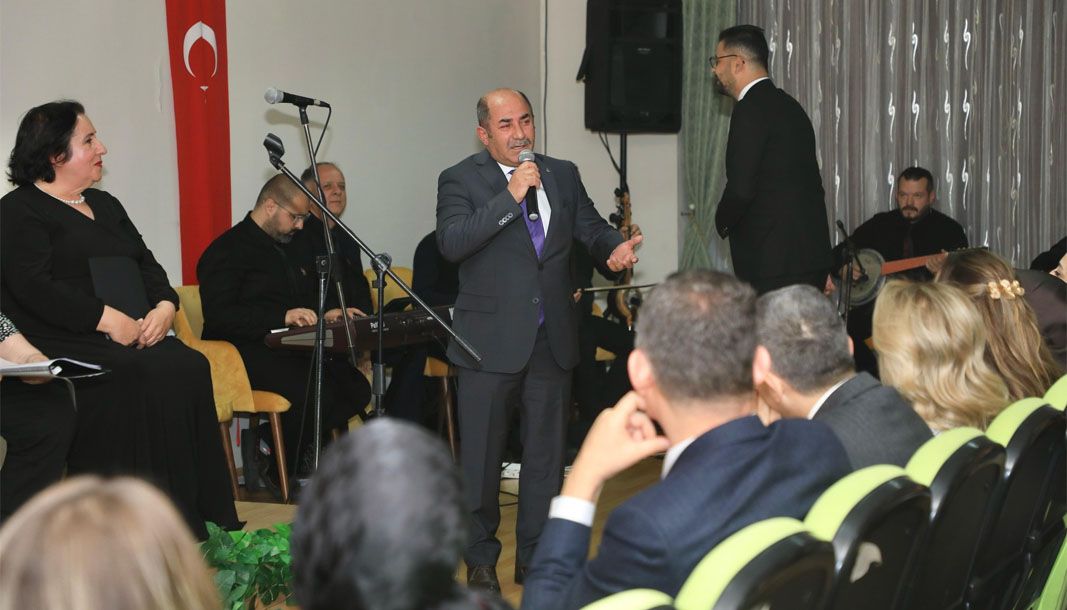 Bir kişi mikrofonla konuşuyor, arka planda müzisyenler ve huzurevi sakinleri oturuyor.