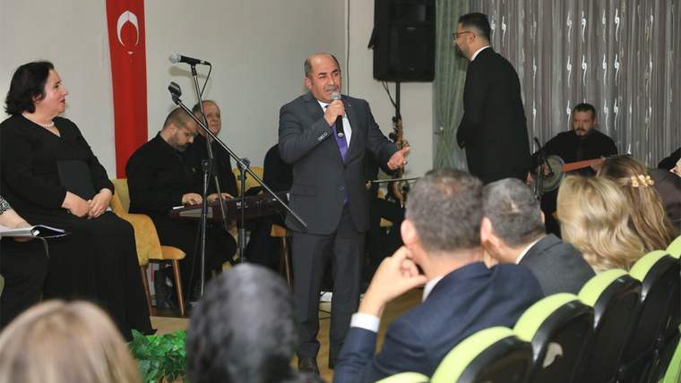 Bir kişi mikrofonla konuşuyor, arka planda müzisyenler ve huzurevi sakinleri oturuyor.