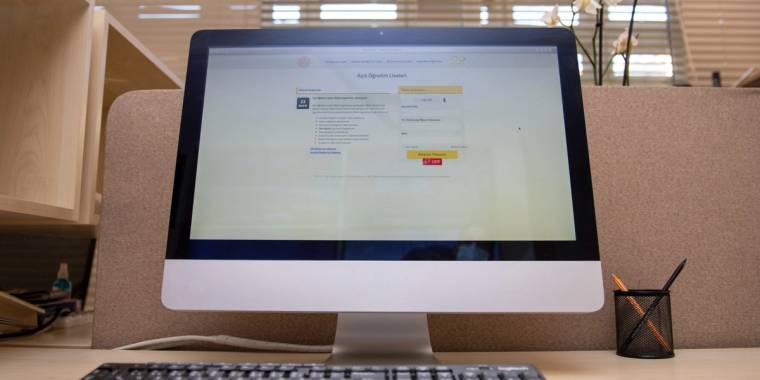 Çalışma masasındaki bilgisayar monitöründe bir web sayfası ve kırmızı bir düğme, yanında kalemlik görülüyor.