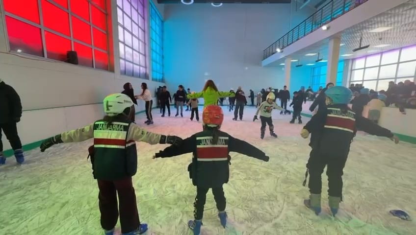 Buz pistinde kayarak eğlenen çocuklar, arka planda kalabalık bir grup yer alıyor.