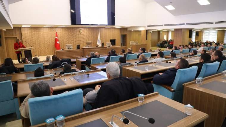 Toplantı salonunda çok sayıda kişi, masalardaki mikrofon ve su şişeleriyle kürsüye bakıyor.
