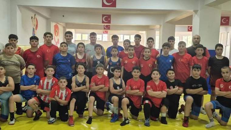 Spor salonunda, kırmızı ve mavi tişörtler giyen çocuklar iki sıra halinde poz veriyor; arkada Türk bayrakları bulunuyor.