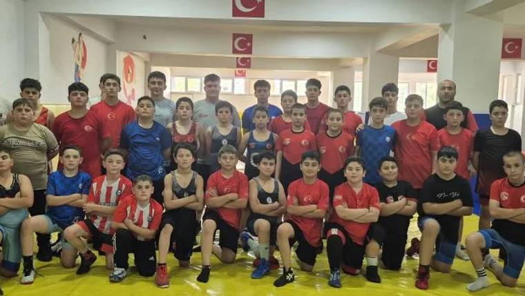 Spor salonunda, kırmızı ve mavi tişörtler giyen çocuklar iki sıra halinde poz veriyor; arkada Türk bayrakları bulunuyor.