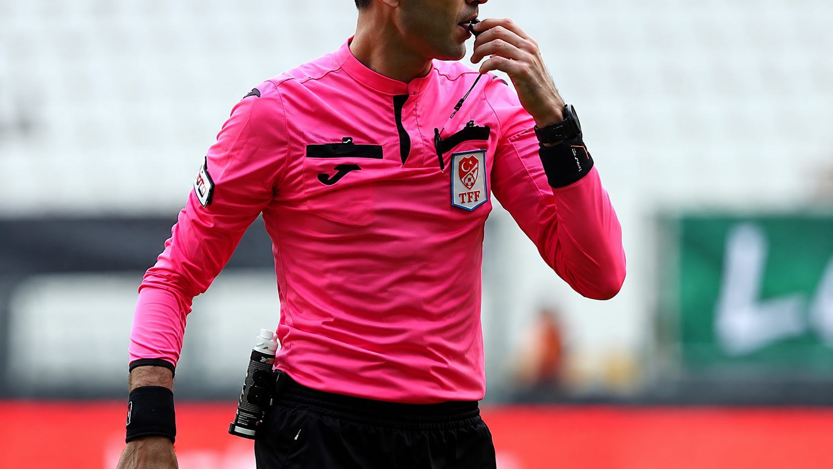 Pembe forma giyen bir futbol hakemi, düdüğünü ağzına götürürken saha içinde görülüyor.
