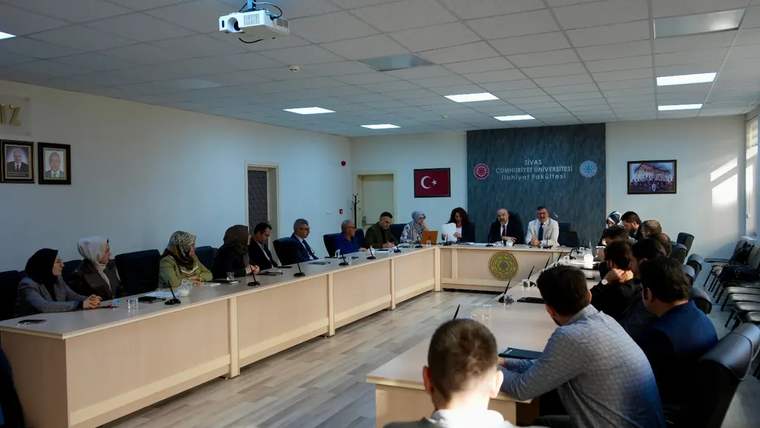 Toplantı salonunda çok sayıda kişi, U biçimindeki masanın çevresinde otururken görülüyor; duvarda Türk bayrağı bulunuyor.