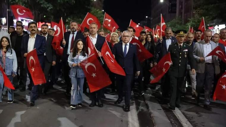 Gece bir caddede Türk bayrakları taşıyan kalabalık arasında sivil ve üniformalı kişiler yürüyor.