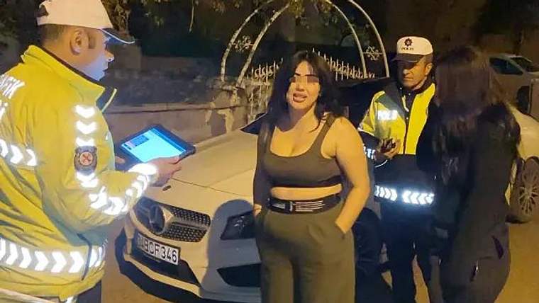 Gece saatlerinde reflektörlü üniformalı iki görevli, beyaz otomobilin yanında bir kadınla konuşuyor.