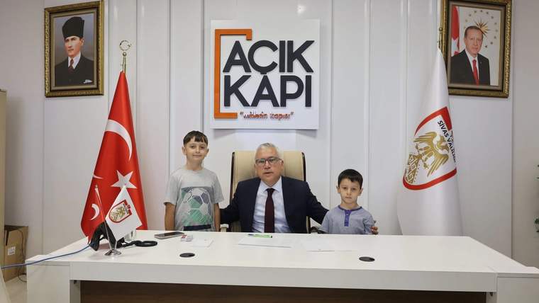Açık Kapı tabelasının önünde, masada oturan bir kişi ve yanında duran iki çocuk görülüyor.