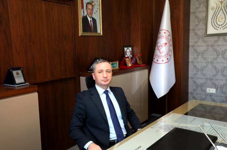 Makam odasında takım elbiseli bir kişi masada oturuyor; arkasında Türk bayrağı ve çerçeveli fotoğraflar bulunuyor.