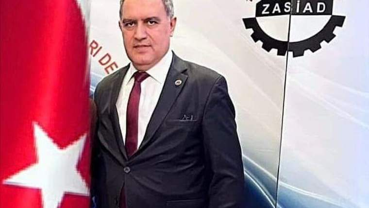 ZASİAD logosunun arkasında, takım elbise giymiş bir kişi, Türk bayrağının yanında duruyor.