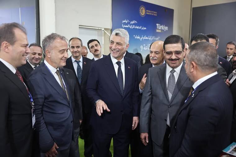 Takım elbiseli bir grup erkek, üzerinde Türkiye yazan mavi bir panonun önünde sohbet ediyor.