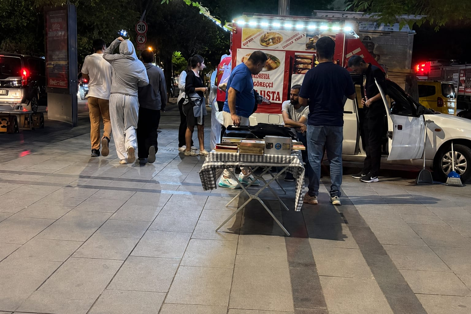 Gece kaldırımında, seyyar yiyecek aracı önünde duran insanlar ve yanında park etmiş beyaz otomobil görülüyor.