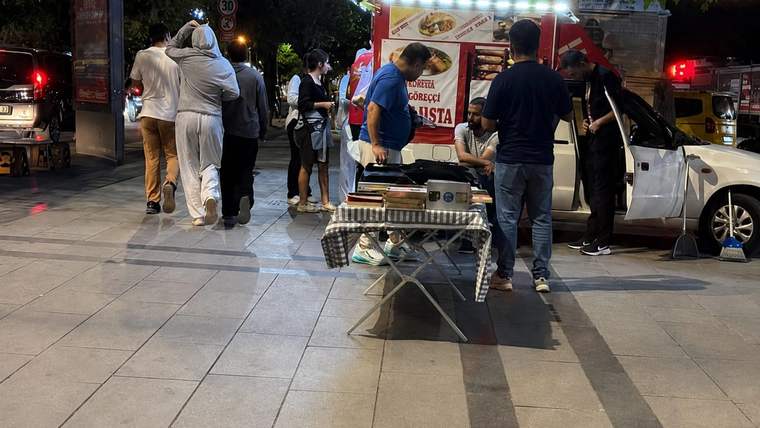 Gece kaldırımında, seyyar yiyecek aracı önünde duran insanlar ve yanında park etmiş beyaz otomobil görülüyor.