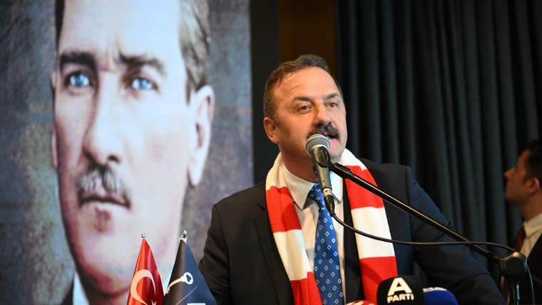 Kürsüde mikrofonla konuşan bir kişi, kırmızı-beyaz atkı ve iki bayrağın önünde duruyor; arkada portre var.
