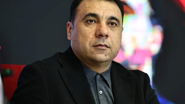 Koyu renk ceket ve gri gömlek giyen bir adam, renkli ve bulanık bir arka plan önünde oturuyor.