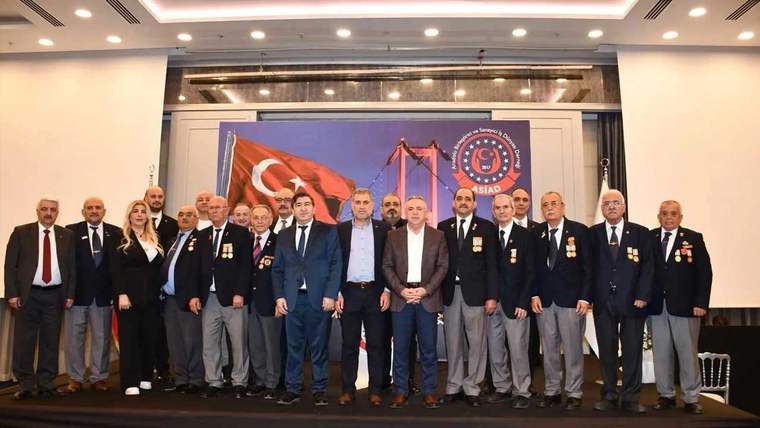 İç mekânda, Türk bayrağı ve amblemlerin bulunduğu bir panonun önünde takım elbiseli grup duruyor.