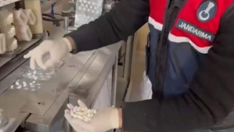 Jandarma yeleği giyen eldivenli bir kişi, metal tezgâhta beyaz kapsül ve tabletleri inceliyor.
