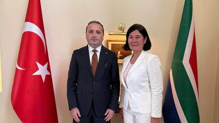 Türk ve Güney Afrika bayrakları arasında takım elbiseli bir erkek ile beyaz kıyafetli bir kadın yan yana duruyor.