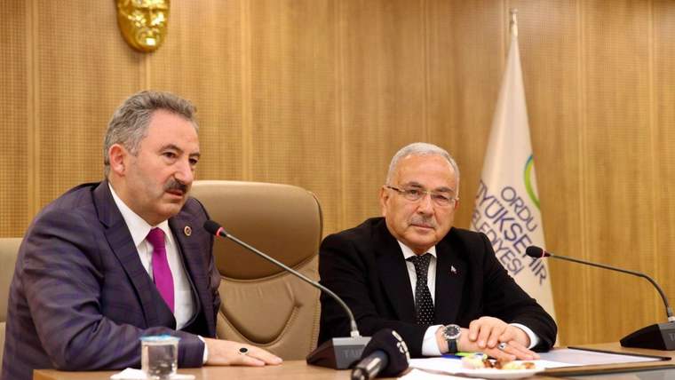 Ahşap panelli salonda mikrofon önünde oturan iki erkek, masadaki belgelerin yanında konuşuyor.