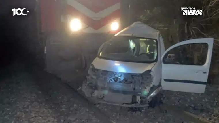 Gece çekiminde, ön kısmı hasarlı beyaz ticari araç tren rayları üzerinde, lokomotifin önünde duruyor.