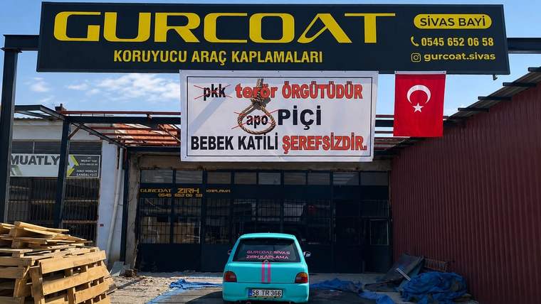 İş yeri önünde siyasi sloganlar yazılı pankart, Türk bayrağı ve park edilmiş turkuaz otomobil görülüyor.