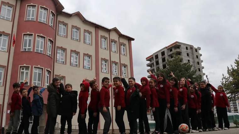 Kırmızı kıyafetler giymiş bir grup öğrenci, okul binasının önünde sırada duruyor.
