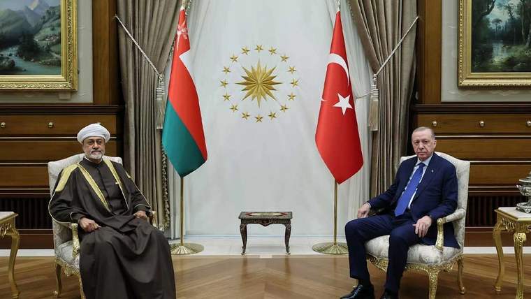 İki kişi, Türk bayrağı ve Umman bayrağının önünde, koltuklarda oturuyor.
