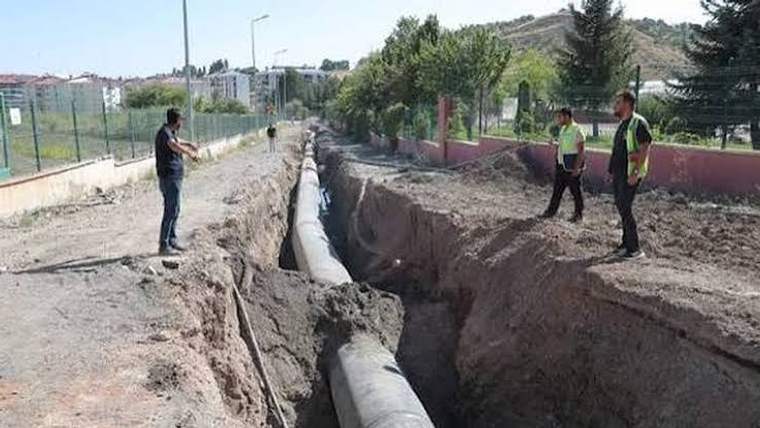 Kazılmış yol boyunca uzanan büyük su borusunun yanında üç kişi çalışma alanında duruyor.
