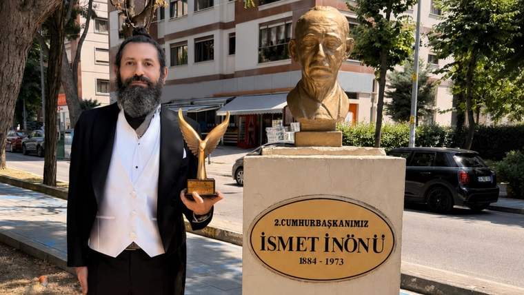 Bir kişi, İsmet İnönü heykelinin önünde, ödül tutarak poz veriyor.