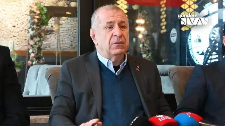 İç mekânda oturan bir adamın önünde kırmızı ve mavi mikrofonlar, yanında iki kişi görülüyor.