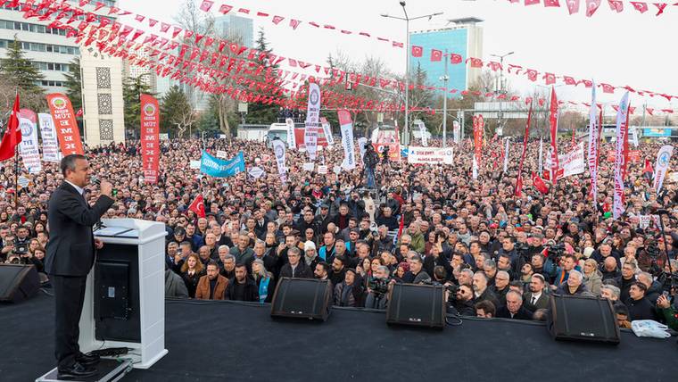 Kürsüde konuşan bir kişi, önündeki kalabalığa seslenirken alanda kırmızı bayraklar ve pankartlar görülüyor.