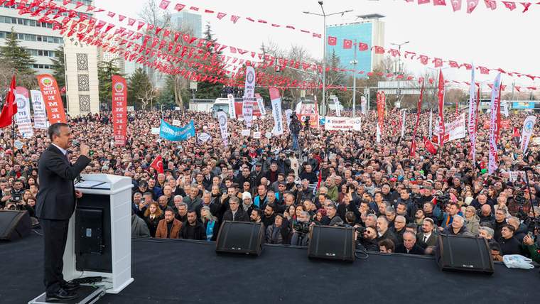 Kürsüde konuşan bir kişi, önündeki geniş kalabalığa seslenirken alanda Türk bayrakları ve flamalar görülüyor.