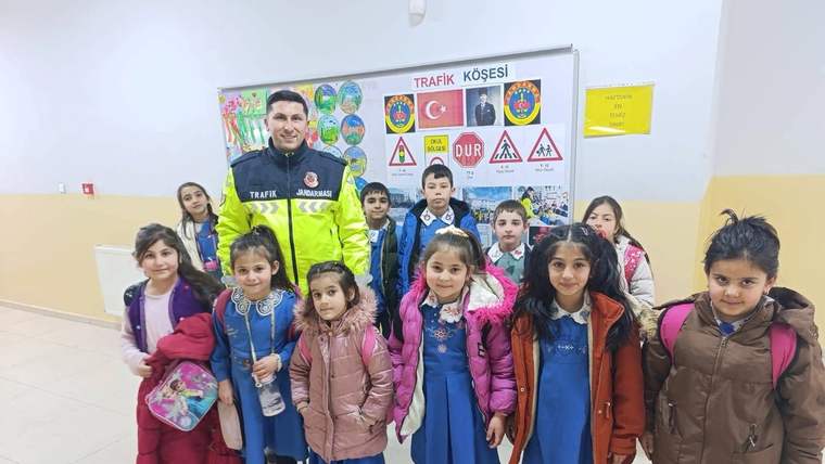 Trafik köşesi panosu önünde, üniformalı bir yetişkin ve okul çağındaki çocuklar birlikte duruyor.