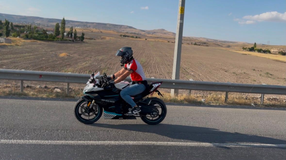 Kasklı bir kişi, kırmızı tişörtle motosiklet üzerinde kırsal yolda ilerlerken görülüyor.